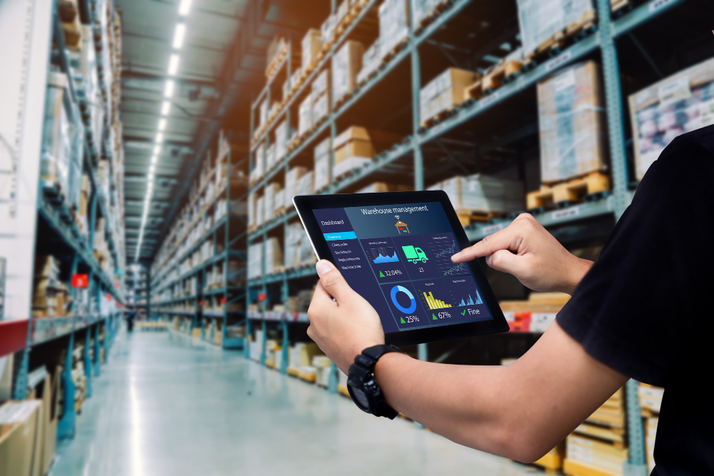 Operator memantau performa gudang melalui tablet digital di dalam smart warehouse modern yang menggunakan teknologi otomatisasi dan IoT.